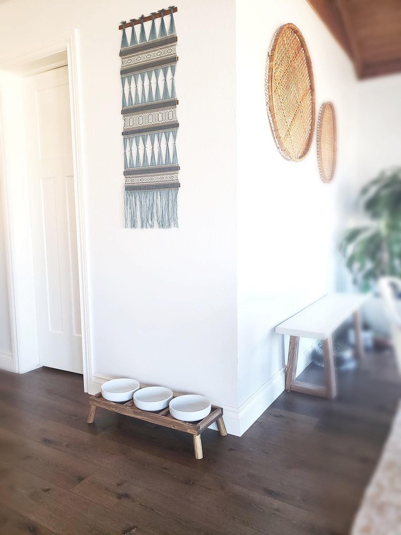 Puede incluir: Un soporte de madera para comida de mascotas con tres cuencos de cer&aacute;mica blanca. El soporte tiene cuatro patas y est&aacute; en una habitaci&oacute;n con suelo de madera dura. Hay un tapiz de pared tejido y una cesta tejida en la pared detr&aacute;s del soporte.
