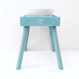 May include: A light blue wooden pet feeder stand with four angled legs and a square top frame. A white heart outline is painted on the front center of the frame. A white ceramic bowl is set into the top opening of the wooden stand.