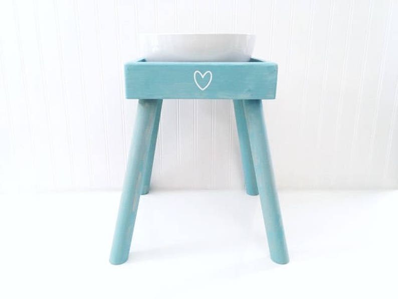 May include: A light blue wooden pet feeder stand with four angled legs and a square top frame. A white heart outline is painted on the front center of the frame. A white ceramic bowl is set into the top opening of the wooden stand.