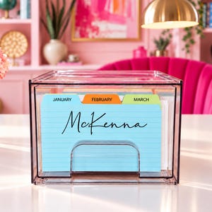May include: A clear acrylic desktop file organizer containing a set of tabbed index cards. The front card is light blue with horizontal lines and the name McKenna printed in black script. The tabs are labeled January, February, and March.