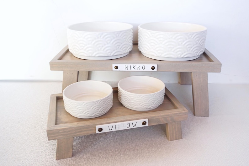 May include: Two elevated dog bowls with a white ceramic bowl with a wave pattern on each. The top level is labelled "NIKKO" and the bottom level is labelled "WILLOW".