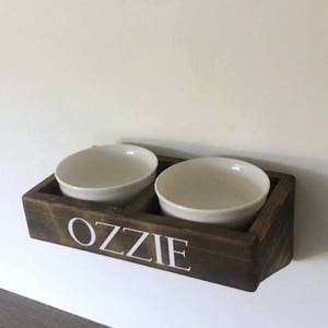 Puede incluir: Un soporte de madera para cuencos de comida para mascotas con dos cuencos de cer&aacute;mica blanca. El soporte est&aacute; te&ntilde;ido de marr&oacute;n oscuro y tiene el nombre "Ozzie" escrito en letras blancas.