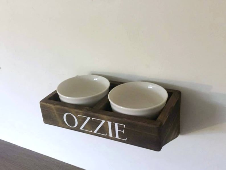 Puede incluir: Un soporte de madera para cuencos de comida para mascotas con dos cuencos de cer&aacute;mica blanca. El soporte est&aacute; te&ntilde;ido de marr&oacute;n oscuro y tiene el nombre "Ozzie" escrito en letras blancas.