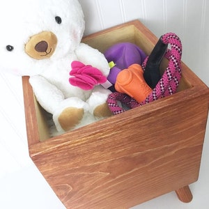 May include: A wooden storage box with a light brown stain, featuring a white teddy bear, a purple toy, an orange chew toy, and a pink rope toy. The box has four short legs.