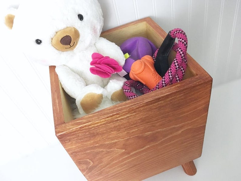 May include: A wooden storage box with a light brown stain, featuring a white teddy bear, a purple toy, an orange chew toy, and a pink rope toy. The box has four short legs.