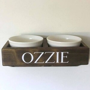 Puede incluir: Un soporte para cuencos de comida para mascotas de madera marr&oacute;n oscuro con el nombre "Ozzie" escrito en letras blancas. Dos cuencos de cer&aacute;mica blanca se colocan en el soporte.