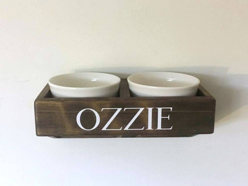 Puede incluir: Un soporte para cuencos de comida para mascotas de madera marr&oacute;n oscuro con el nombre "Ozzie" escrito en letras blancas. Dos cuencos de cer&aacute;mica blanca se colocan en el soporte.