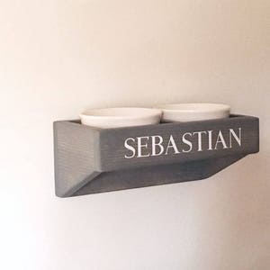 Puede incluir: Un soporte de cuencos para comida de mascotas de madera gris con el nombre "Sebastian" en la parte delantera. Dos cuencos de cer&aacute;mica blanca est&aacute;n montados en el soporte.
