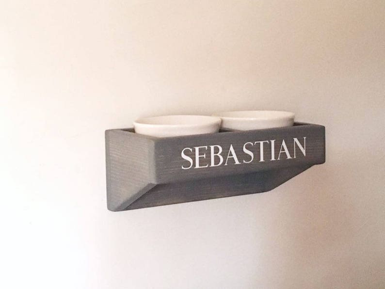 Puede incluir: Un soporte de cuencos para comida de mascotas de madera gris con el nombre "Sebastian" en la parte delantera. Dos cuencos de cer&aacute;mica blanca est&aacute;n montados en el soporte.