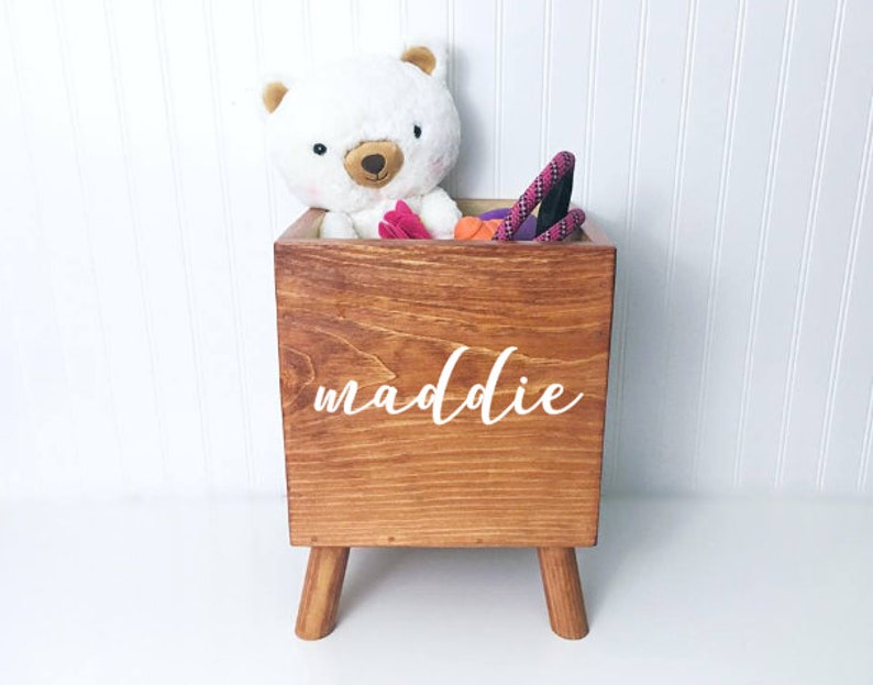 May include: A wooden toy storage box with four legs, painted in a dark brown stain. The box has a white inscription that reads "maddie". The box is filled with toys, including a white stuffed bear, a pink and purple rope, and a red and orange rope.