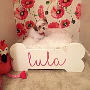 May include: A small dog lies on a white, bone-shaped dog bed with the name "Lula" in pink script. The bed is covered with a fluffy white material. A pink flamingo toy is on the left, and floral wallpaper is in the background.