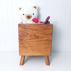 May include: A wooden toy storage box with four legs. The box is stained a dark brown color and has a white teddy bear and pink and orange toys inside.