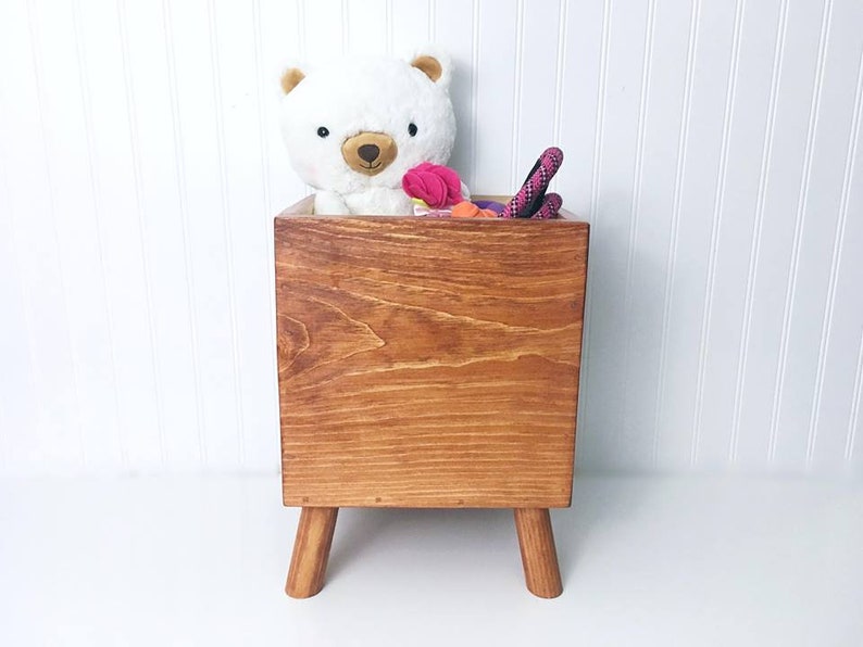 May include: A wooden toy storage box with four legs. The box is stained a dark brown color and has a white teddy bear and pink and orange toys inside.