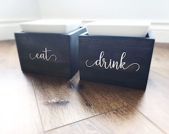 May include: Two black wooden boxes with white ceramic bowls on top. The boxes are labeled "eat" and "drink" in white cursive lettering.