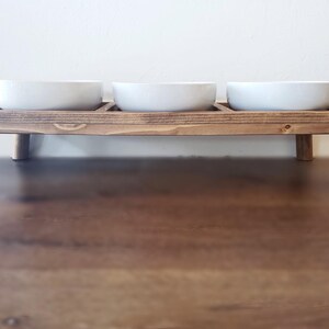 Puede incluir: Un soporte de madera para comida de mascotas con tres cuencos de cer&aacute;mica blanca. El soporte tiene cuatro patas y est&aacute; te&ntilde;ido de un color marr&oacute;n oscuro.