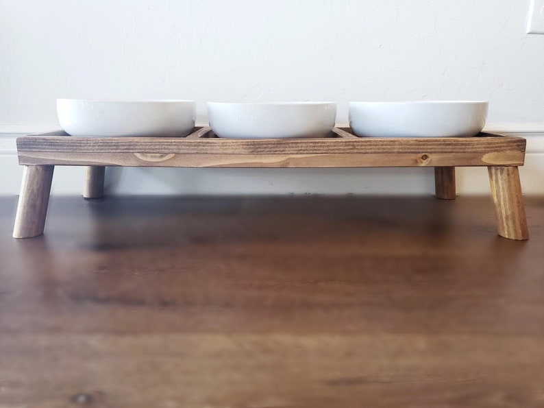 Puede incluir: Un soporte de madera para comida de mascotas con tres cuencos de cer&aacute;mica blanca. El soporte tiene cuatro patas y est&aacute; te&ntilde;ido de un color marr&oacute;n oscuro.
