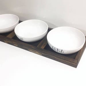 May include: Three white ceramic bowls with black lettering on a dark brown wooden tray. The bowls are labelled "Max", "Ari", and "Luna".
