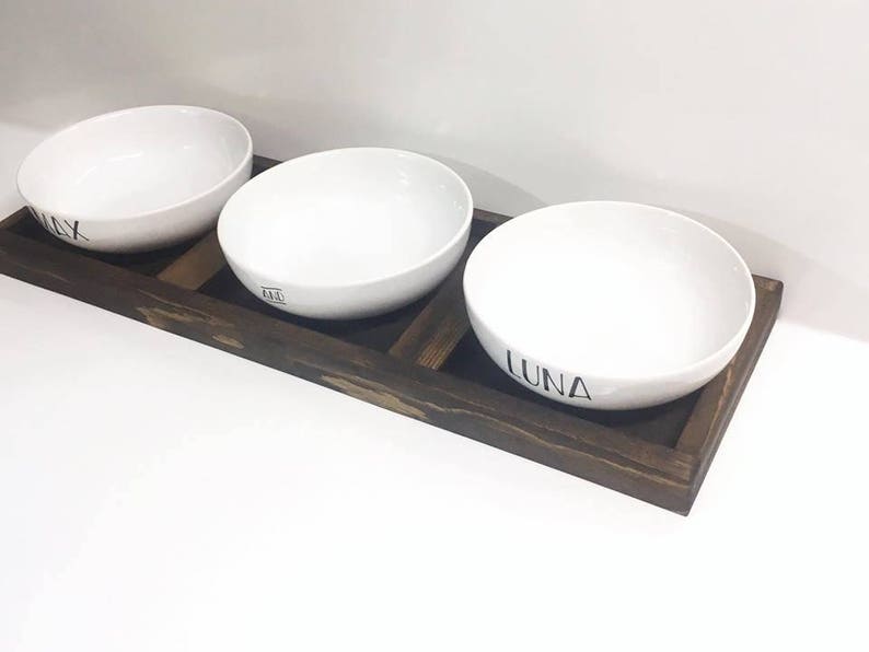 May include: Three white ceramic bowls with black lettering on a dark brown wooden tray. The bowls are labelled "Max", "Ari", and "Luna".