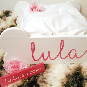 May include: A white, cloud-shaped bed frame with the name "Lula" in pink script. A small pink sign reads "Lula's room." The bed is made with a white pillow and blanket, and decorated with pink flowers. The bed is on a faux fur rug.