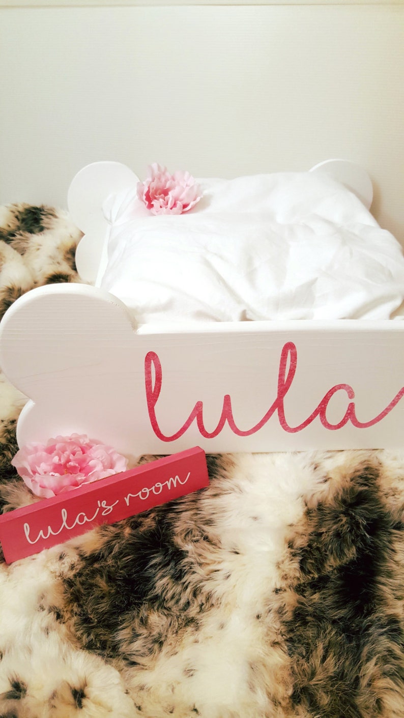 May include: A white, cloud-shaped bed frame with the name "Lula" in pink script. A small pink sign reads "Lula's room." The bed is made with a white pillow and blanket, and decorated with pink flowers. The bed is on a faux fur rug.