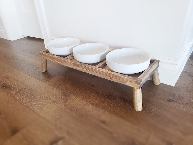 Puede incluir: Un soporte elevado de madera para cuencos de comida y agua para mascotas con tres cuencos de cer&aacute;mica blanca. El soporte tiene cuatro patas y est&aacute; te&ntilde;ido de un color marr&oacute;n oscuro.
