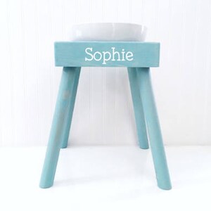 May include: A light blue wooden stool with four angled legs and a rectangular top. The front of the stool features the name Sophie printed in white, sans-serif capital and lowercase letters. A white ceramic bowl sits on the top surface.