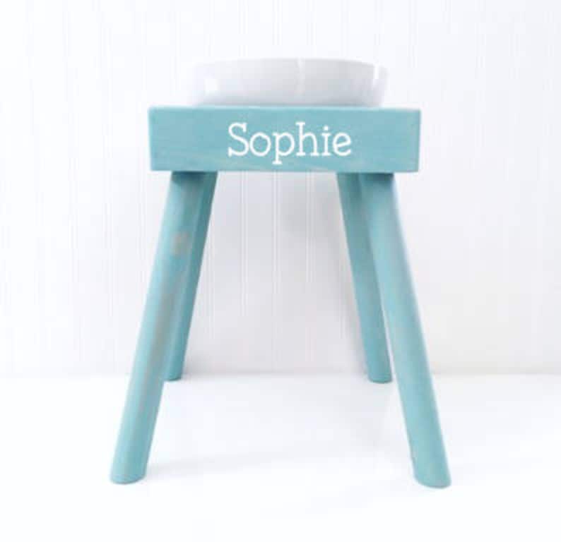 May include: A light blue wooden stool with four angled legs and a rectangular top. The front of the stool features the name Sophie printed in white, sans-serif capital and lowercase letters. A white ceramic bowl sits on the top surface.