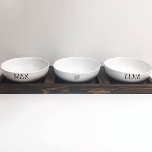 May include: Three white ceramic pet bowls with the names Max, Ziva, and Luna printed on them. The bowls are sitting on a dark brown wooden stand.