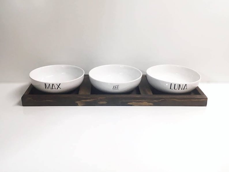 May include: Three white ceramic pet bowls with the names Max, Ziva, and Luna printed on them. The bowls are sitting on a dark brown wooden stand.
