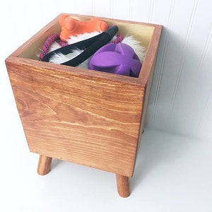 May include: A wooden toy storage box with four legs. The box is filled with dog toys, including a purple ball, a rope toy, and an orange toy.