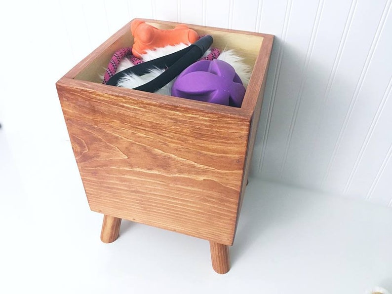 May include: A wooden toy storage box with four legs. The box is filled with dog toys, including a purple ball, a rope toy, and an orange toy.