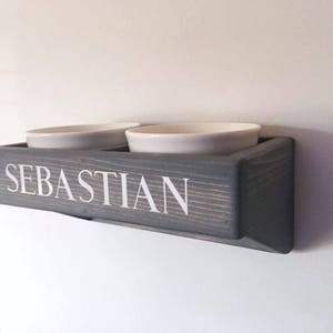 Puede incluir: Un soporte de cuencos para comida de mascotas de madera gris con el nombre "Sebastian" grabado en la parte delantera. Dos cuencos de cer&aacute;mica blanca se colocan en la parte superior del soporte.