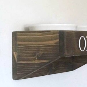Puede incluir: Una estanter&iacute;a de madera marr&oacute;n oscuro con el nombre "OZZIE" escrito en letras blancas. La estanter&iacute;a est&aacute; dise&ntilde;ada para sostener dos cuencos blancos.