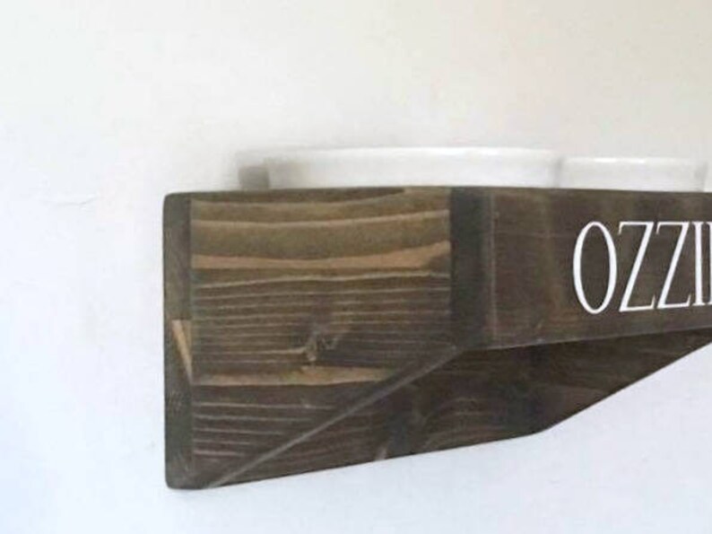 Puede incluir: Una estanter&iacute;a de madera marr&oacute;n oscuro con el nombre "OZZIE" escrito en letras blancas. La estanter&iacute;a est&aacute; dise&ntilde;ada para sostener dos cuencos blancos.