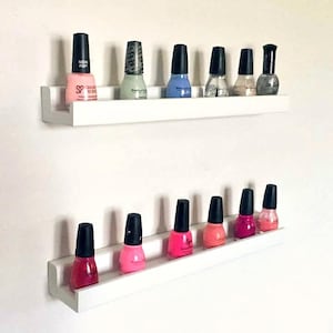 May include: Two white shelves with a variety of nail polish bottles in different colors, including pink, blue, green, and clear. The shelves are mounted on a white wall.
