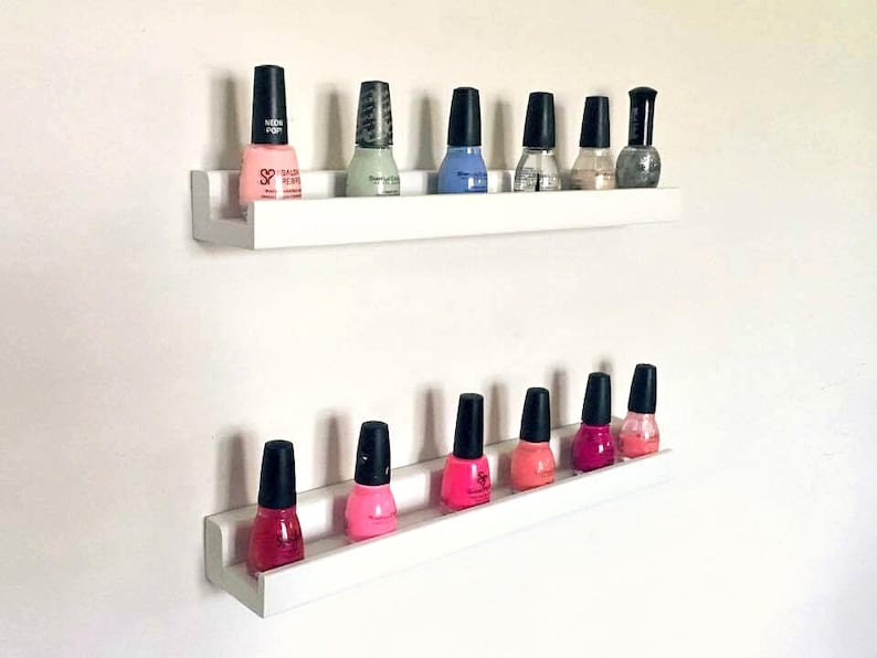 May include: Two white shelves with a variety of nail polish bottles in different colors, including pink, blue, green, and clear. The shelves are mounted on a white wall.
