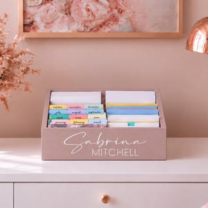 May include: A mauve wooden desktop organizer featuring the name Sabrina Mitchell in white script and block lettering. The interior contains two rows of dividers with colorful tabs labeled with months of the year, holding white and blue cards.