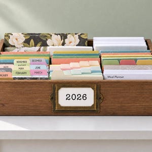 May include: A rectangular wooden storage box with a brass-toned metal label holder on the front containing a white card with the text 2026. The box is filled with organized, multi-colored tabbed index cards and dividers labeled with months.