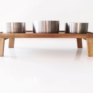 May include: A wooden elevated pet food and water bowl stand with three stainless steel bowls. The stand has four legs and is made of natural wood.