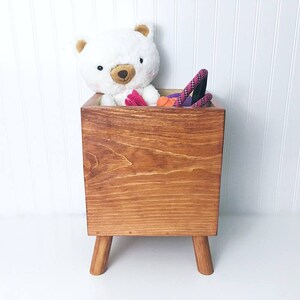 May include: A wooden toy storage box with four legs. The box is filled with toys, including a white teddy bear, a pink rope, and a red and orange toy.