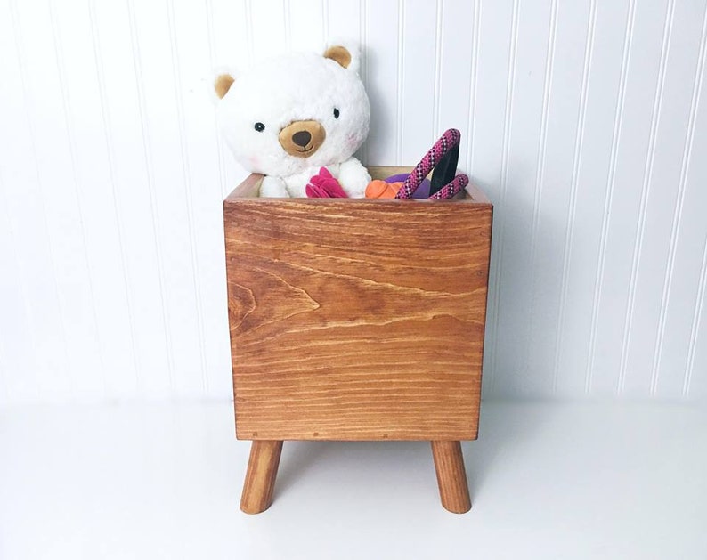 May include: A wooden toy storage box with four legs. The box is filled with toys, including a white teddy bear, a pink rope, and a red and orange toy.