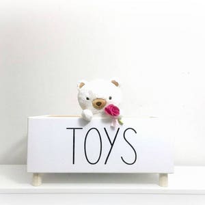 May include: White wooden toy box with the word "TOYS" printed on the front. A white stuffed bear with a pink flower is peeking out of the box.