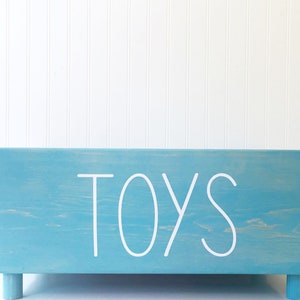 May include: A light blue wooden toy box with the word "TOYS" written in white on the front. The box has four small legs.