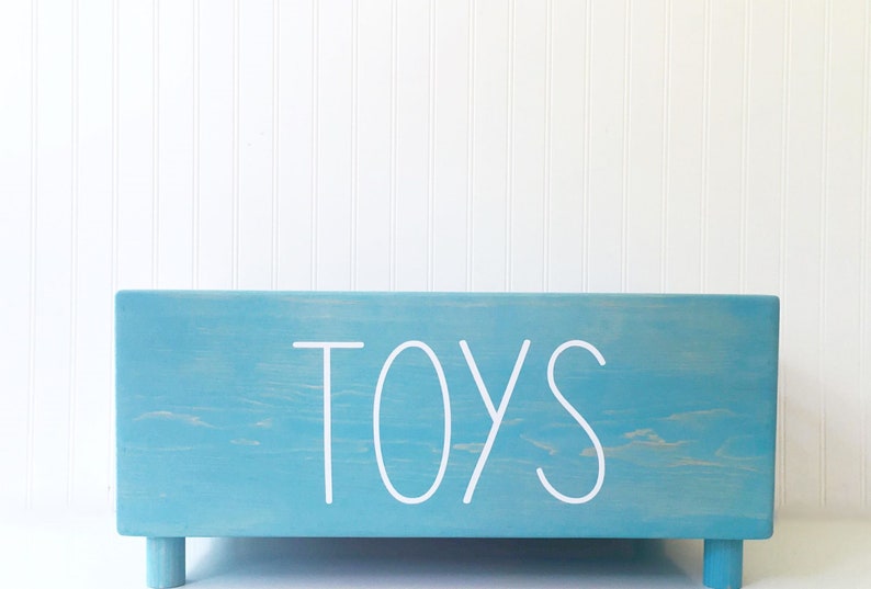 May include: A light blue wooden toy box with the word "TOYS" written in white on the front. The box has four small legs.