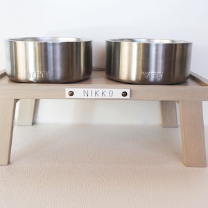 May include: Two stainless steel dog bowls with the word "YETI" engraved on them, sitting on a light wood stand with the name "NIKKO" engraved on a white plaque.
