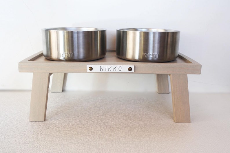 May include: Two stainless steel dog bowls with the word "YETI" engraved on them, sitting on a light wood stand with the name "NIKKO" engraved on a white plaque.