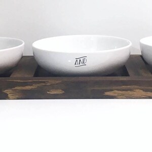 May include: Three white ceramic pet bowls with the names Max, And, and Luna printed on them, sitting on a dark brown wooden tray.