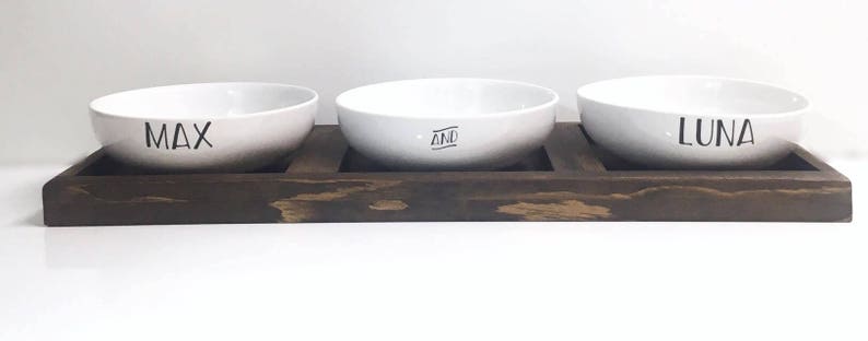 May include: Three white ceramic pet bowls with the names Max, And, and Luna printed on them, sitting on a dark brown wooden tray.