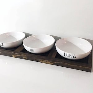 May include: Three white ceramic bowls with the names "Max", "Zia", and "Luna" printed on them, sitting on a dark brown wooden tray.