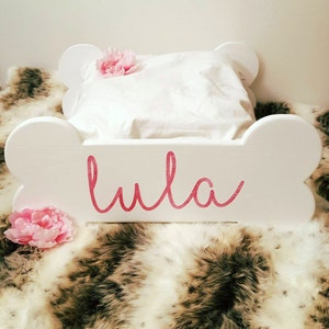 May include: A white, bone-shaped dog bed with the name "Lula" in pink script. It features a white pillow and is adorned with pink flowers. The bed rests on a faux fur blanket.